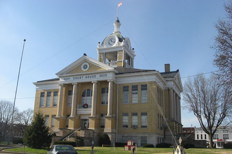 The yellow brick Warrick County Courthouse with a columned front and a clock tower, seen across the courthouse lawn