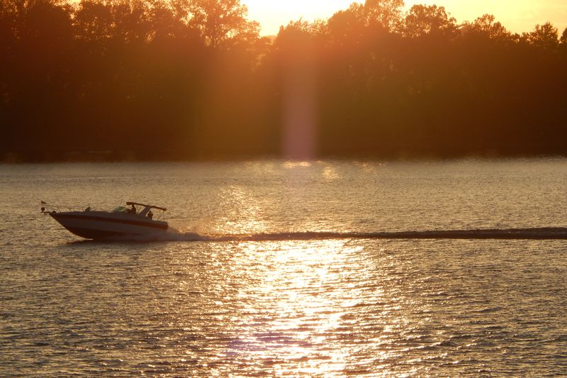 A motorboat crossing the Ohio River at sunset, with the tree-lined far bank in silhouette